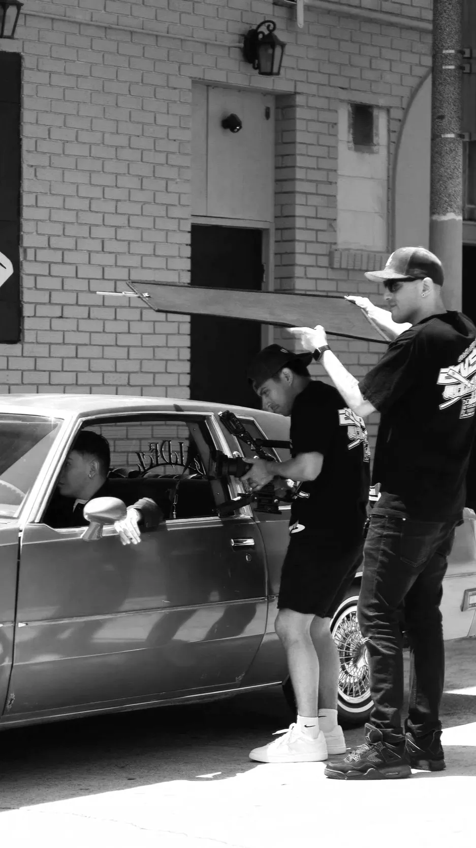 Black and white photo of filmmakers shooting a scene with an actor in a car on a city street.
