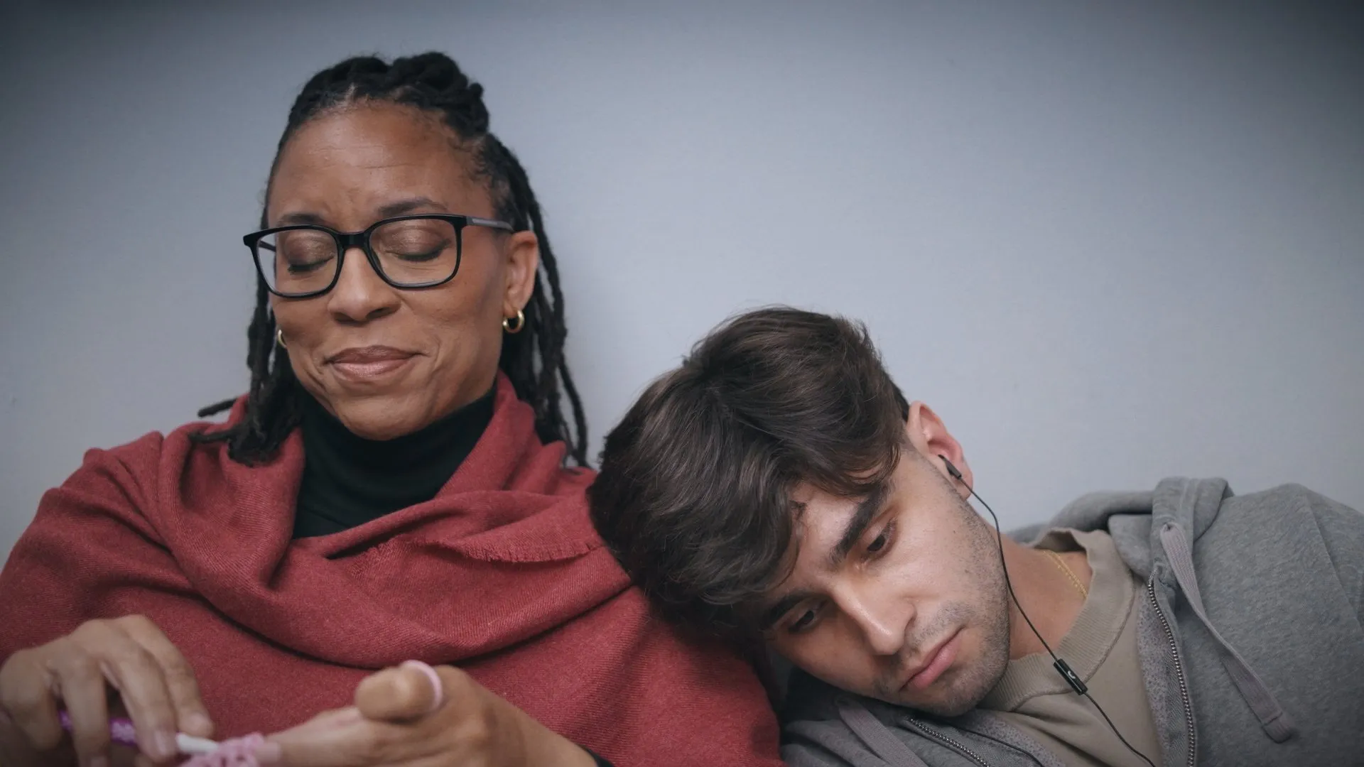 A woman in glasses and a red shawl smiles softly as a young man rests his head on her shoulder.