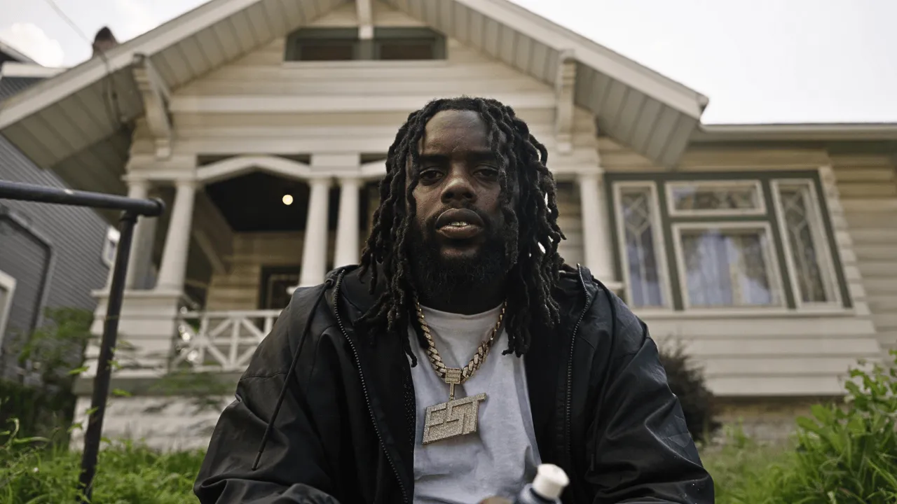 Man in a black jacket and chain standing in front of a white wooden house, filmed for The Taboo Room.