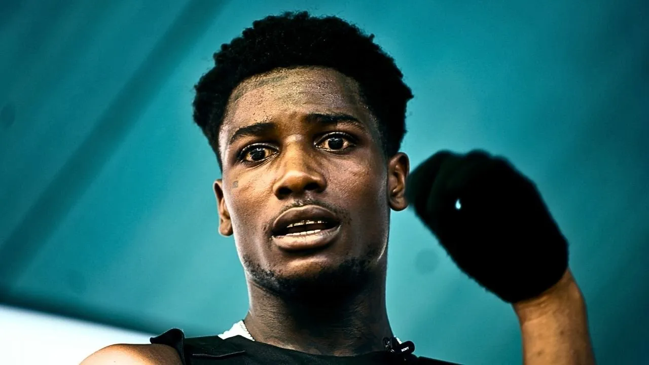 Young man in a black vest against a teal background, mid-conversation.