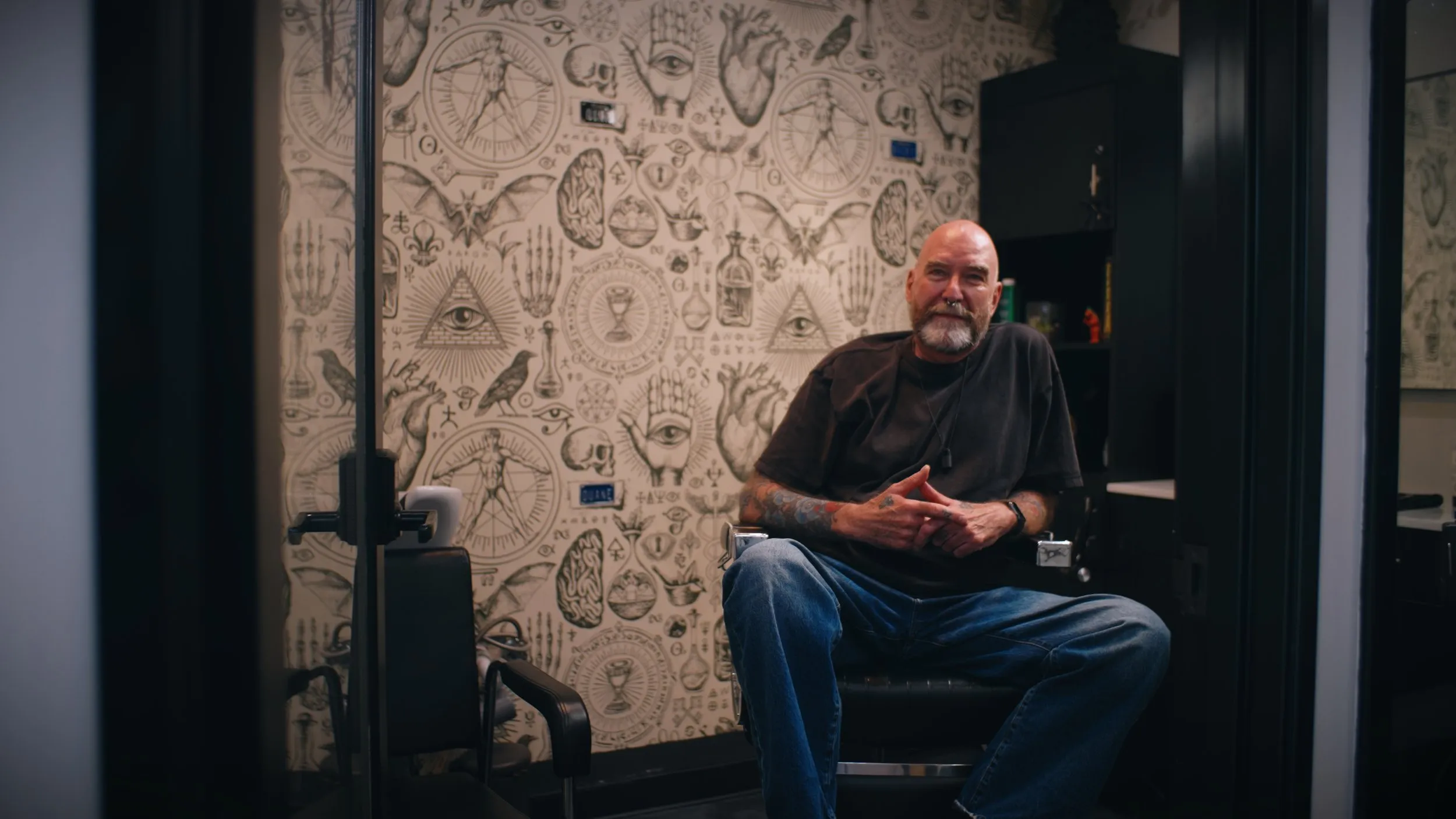 Barber with a white beard seated in his suite in front of tattoo-flash wallpaper.
