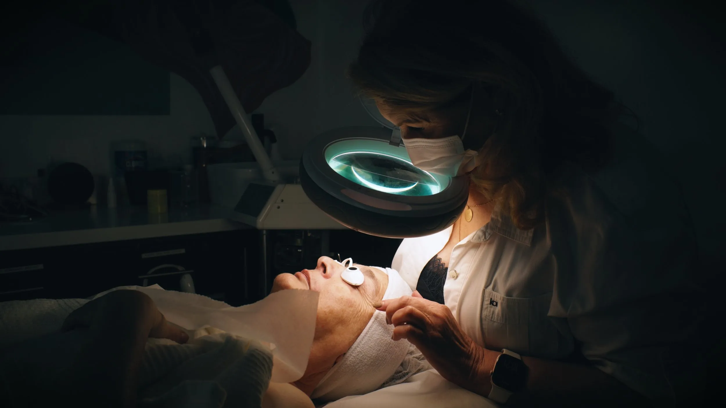 Esthetician under a magnifying lamp performing a facial treatment on a client.