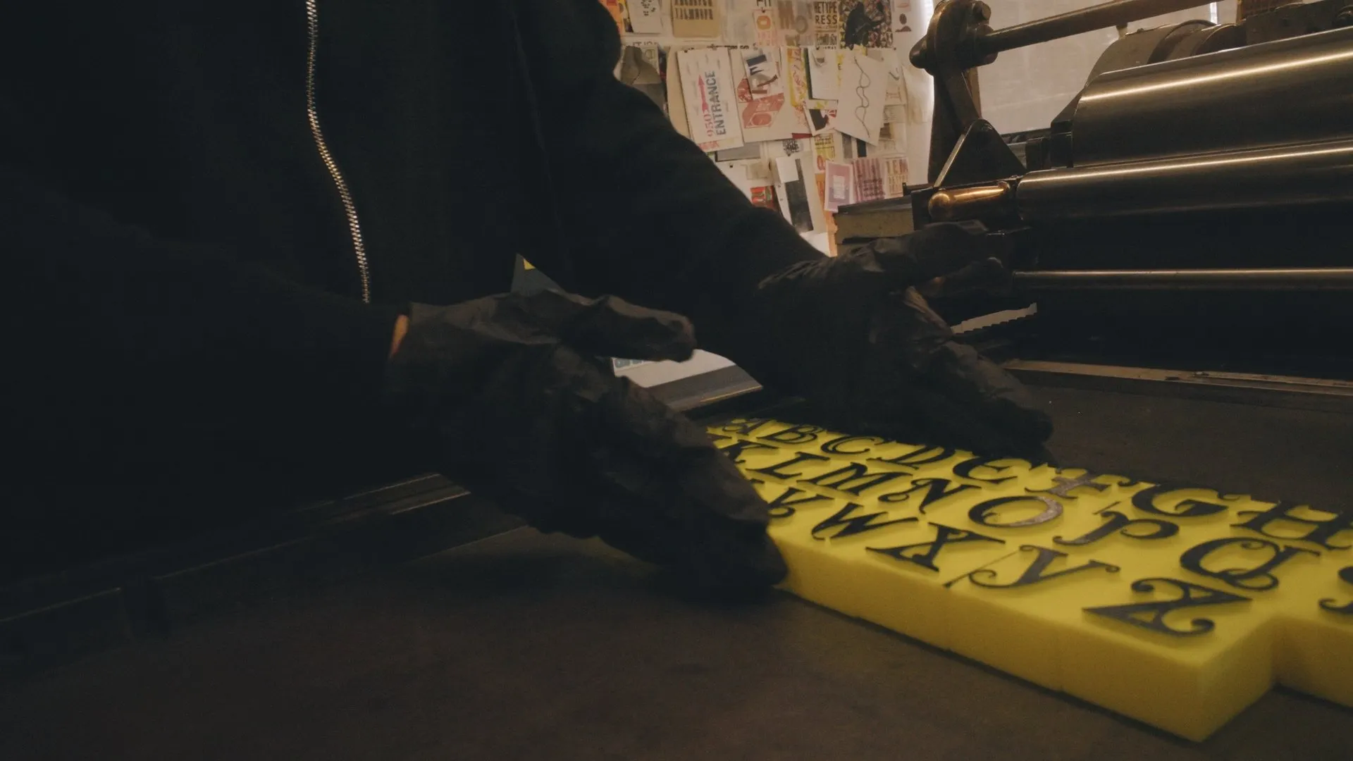 Gloved hands setting yellow letterpress type on a dark table.